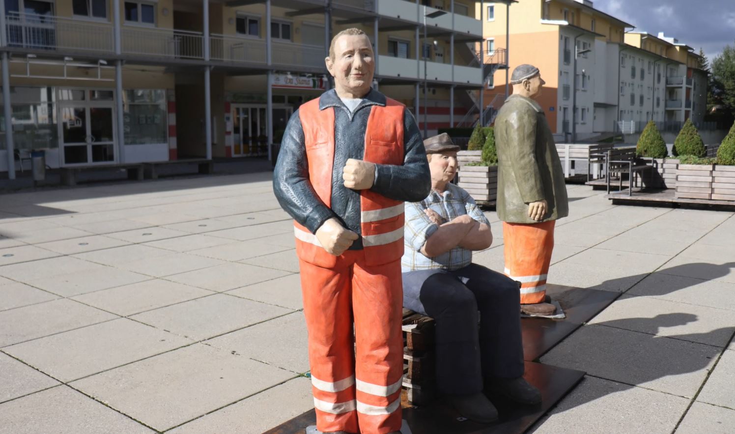 MABP Außenbereich mit orangefarbigen Skulpturen in Schutzkleidung, drei Figuren als Kunstinstallation vor Mehrfamilienhäusern auf dem Platz