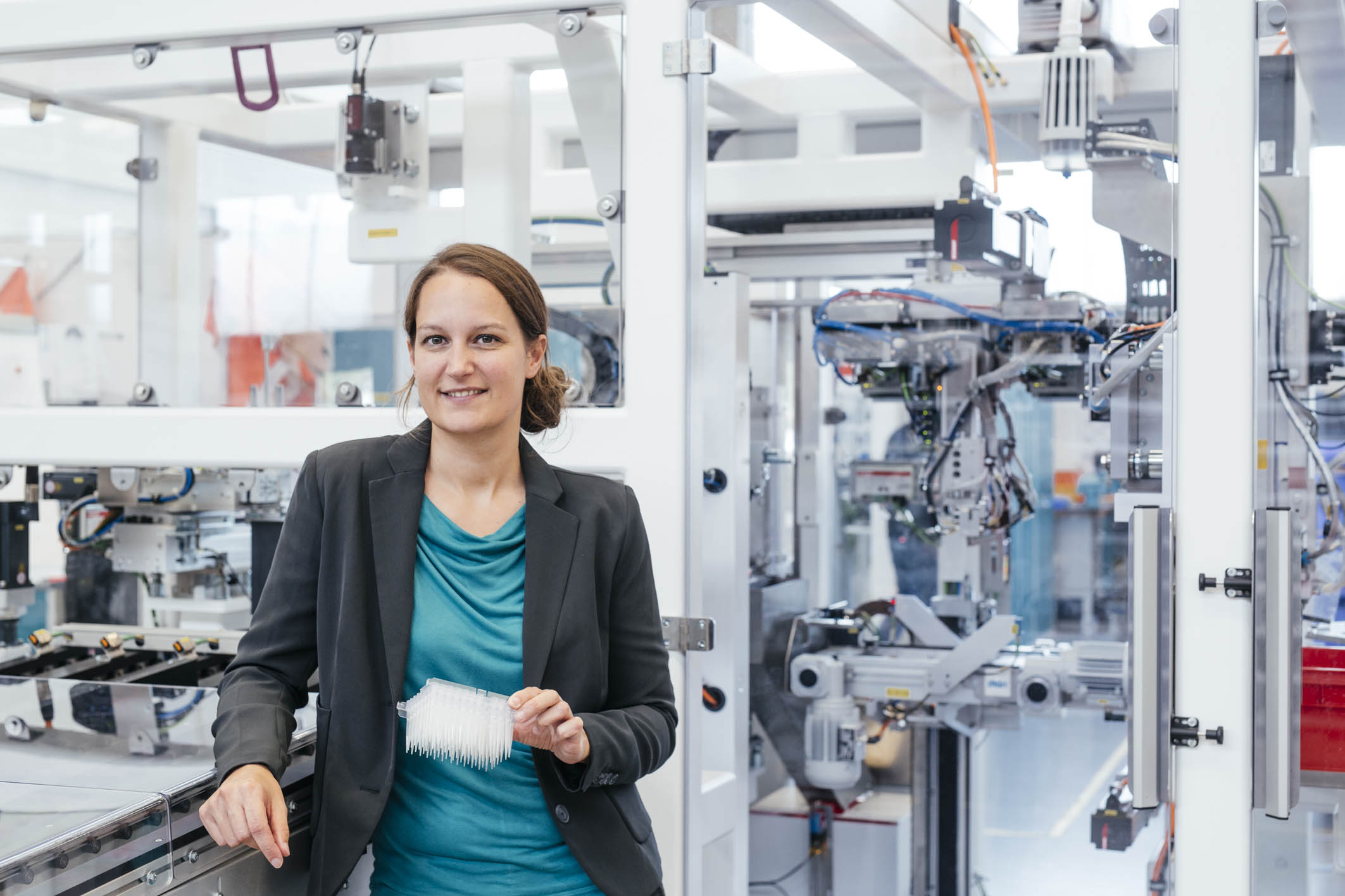 Quirin Leppert in a blue-green blouse and black blazer in an industrial production hall with an automated assembly line at the MABP Munich Airport Business Park site