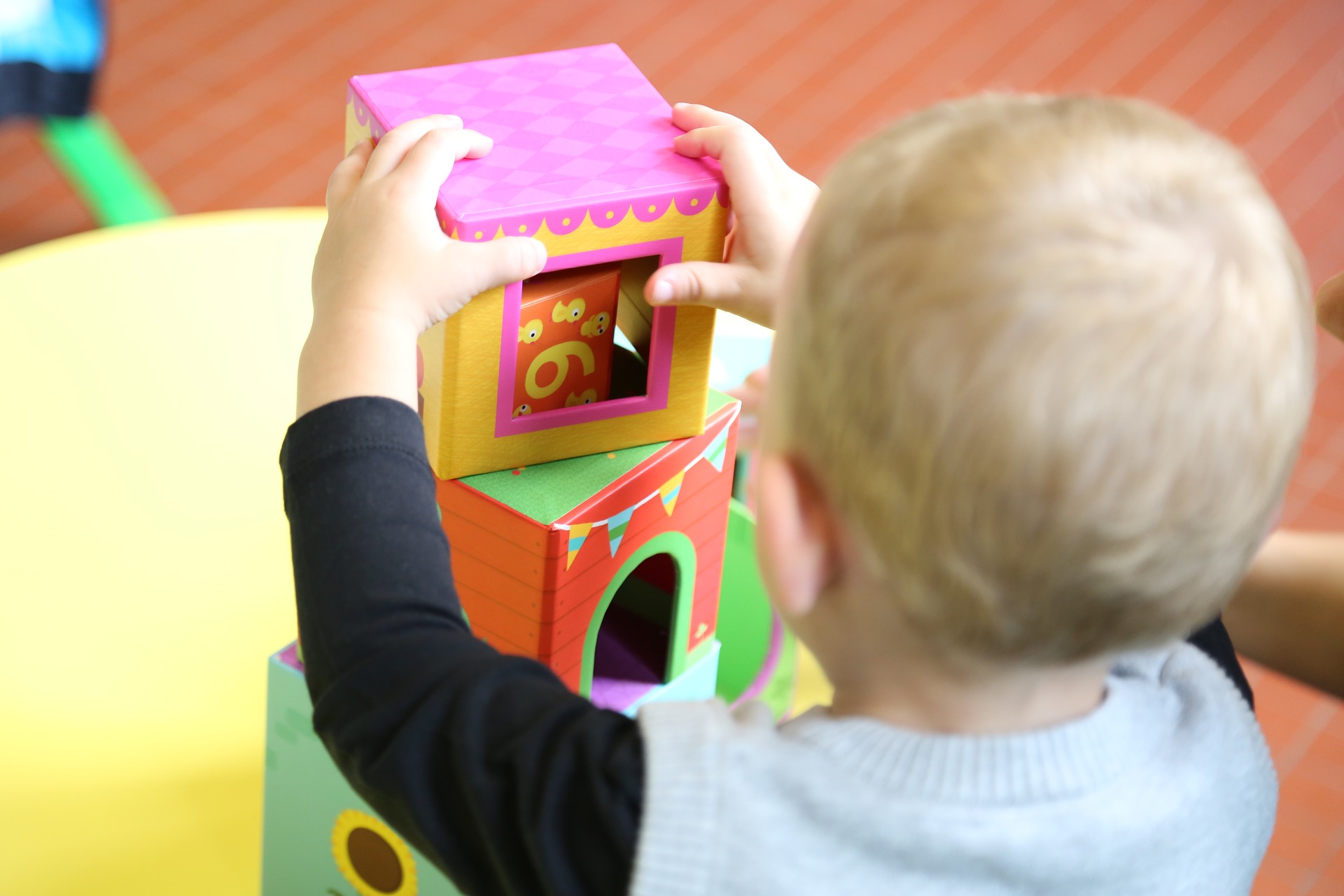 Colorful cardboard stacking blocks are stacked by a child; pink cube on yellow block, green accents, red base in the background.