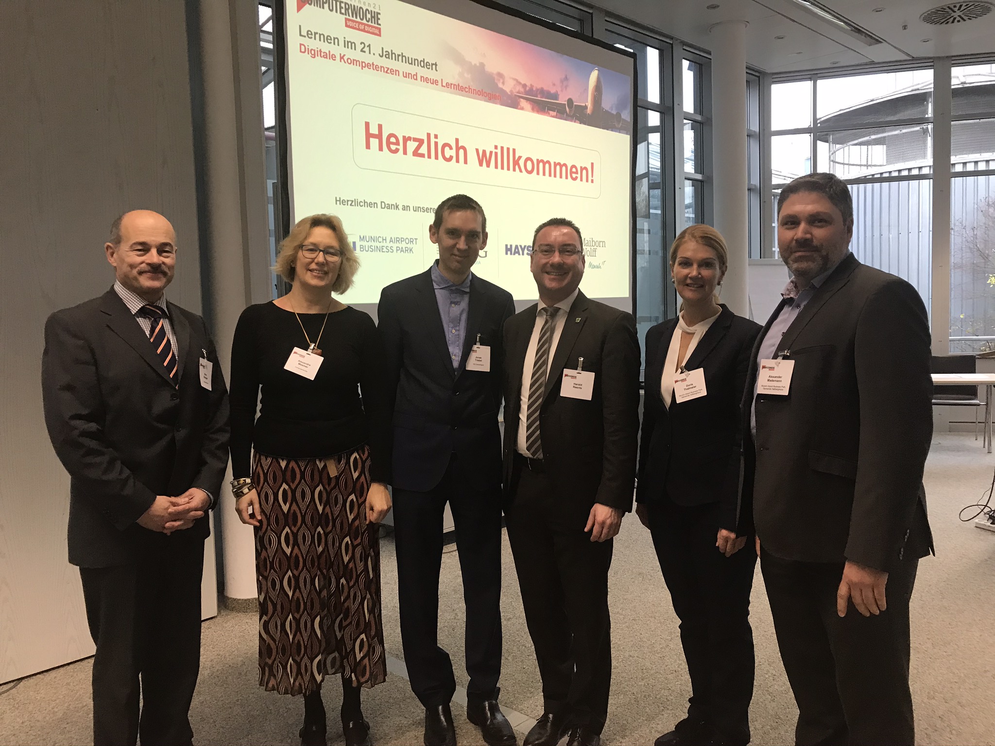 Gruppe von sechs Personen in formeller Kleidung bei einer Computerwoche-Tagung, helles Veranstaltungszentrum, Namensschilder, professionelle Business-Atmosphäre