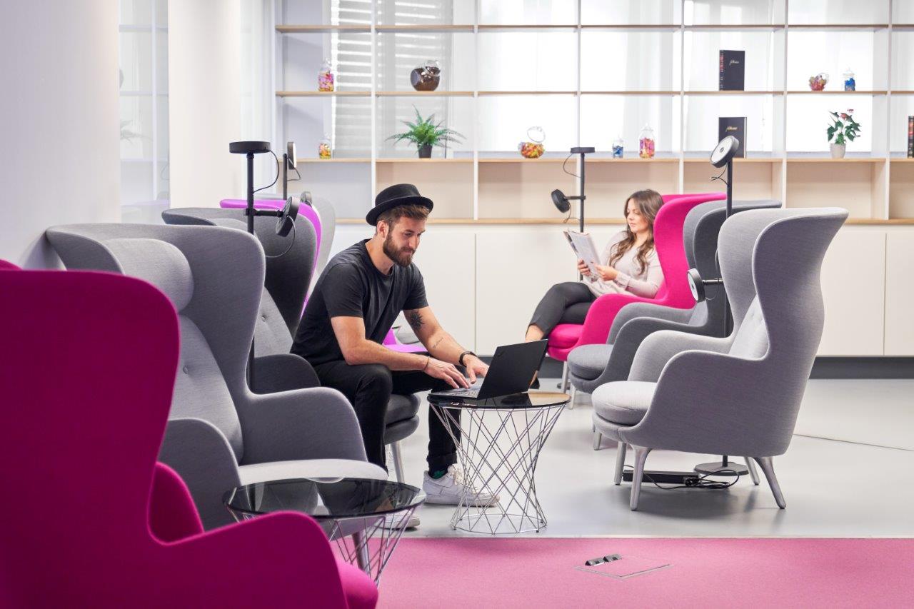 SiriusFacilities Lounge with pink and gray armchairs, a man with a laptop, a woman sitting in the background reading in a bright room.