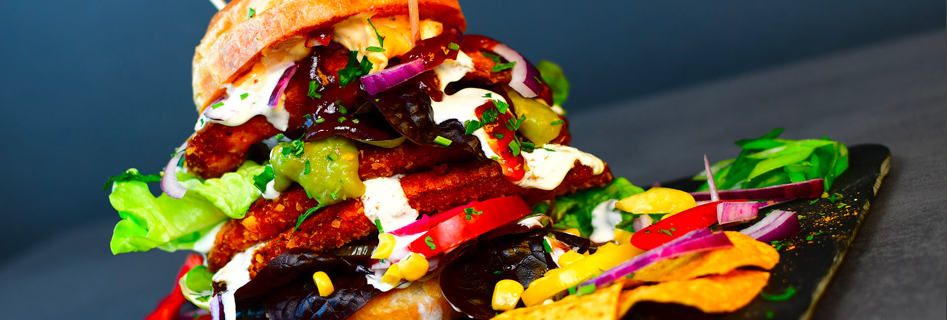 Tasty Gorilla Burger auf einem Holzbrett, bunt belegt mit Salat, Käse, roten Zwiebeln, Sauce und Mais – ein farbenfrohes, satt wirkendes Food-Foto.