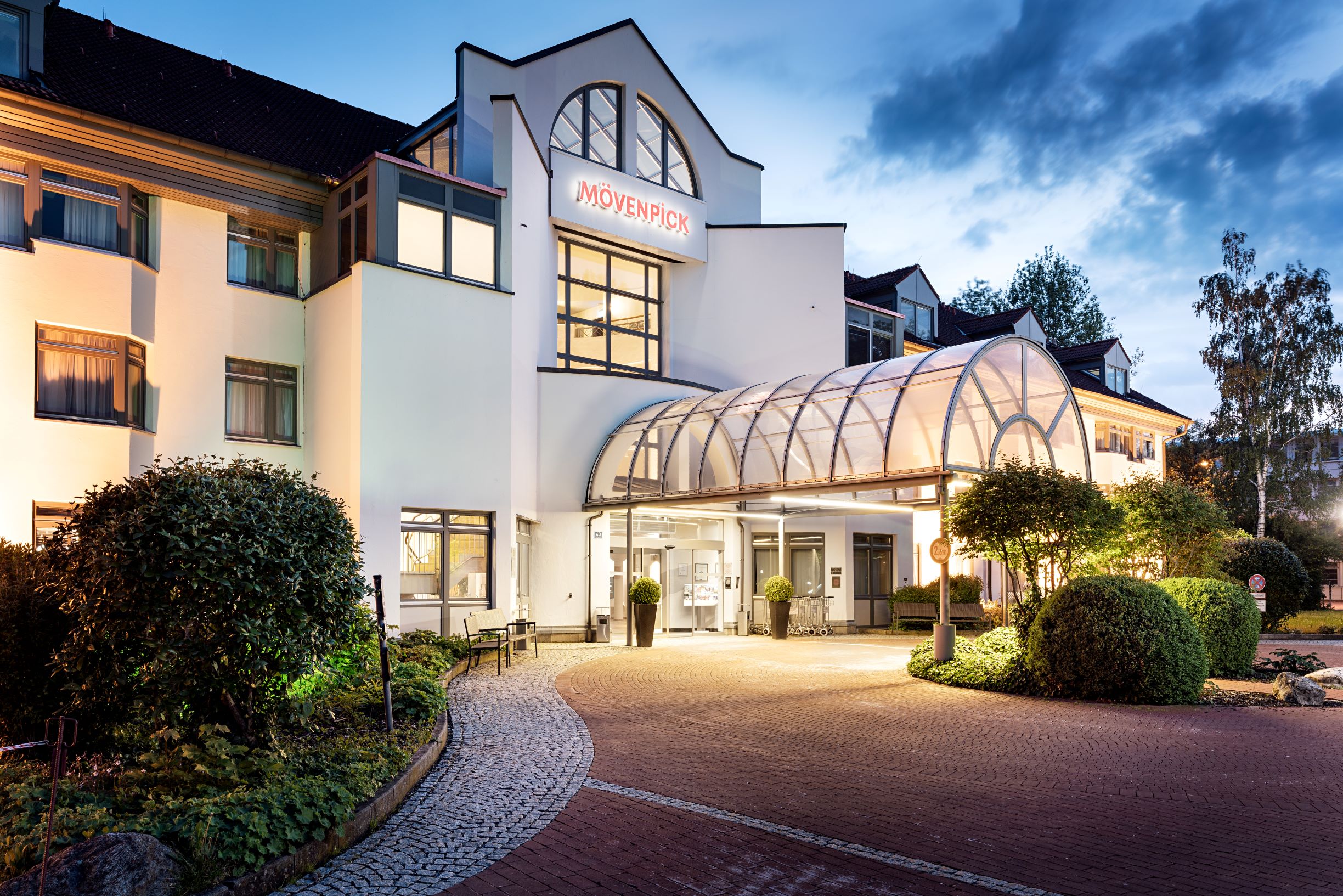 Mövenpick Hotel Eingang mit gläsernem Vordach, beleuchtete Fassade und gepflegter Außenbereich am Abend; Hotellogo sichtbar, Zugang in der Mitte, umliegende Bäume und Pflanzkübel.
