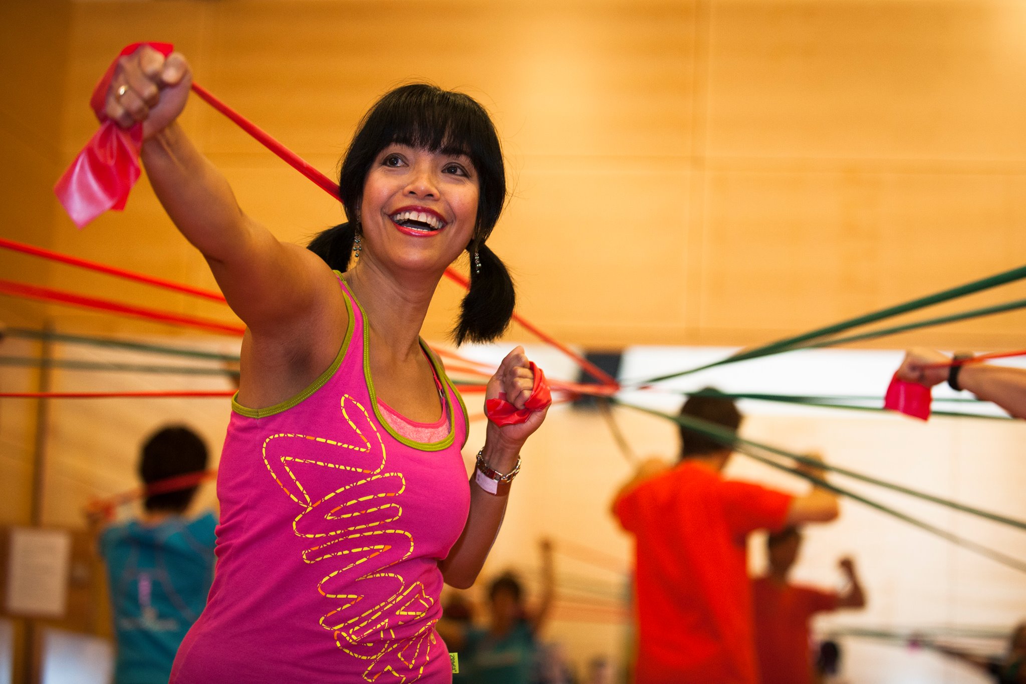 Person in pinkem Fitnesstop mit gelbem Muster beim Fitness-Workshop, rotes Band im Arm, gelb-oranger Hintergrund, aktive Gruppenübung im Hintergrund