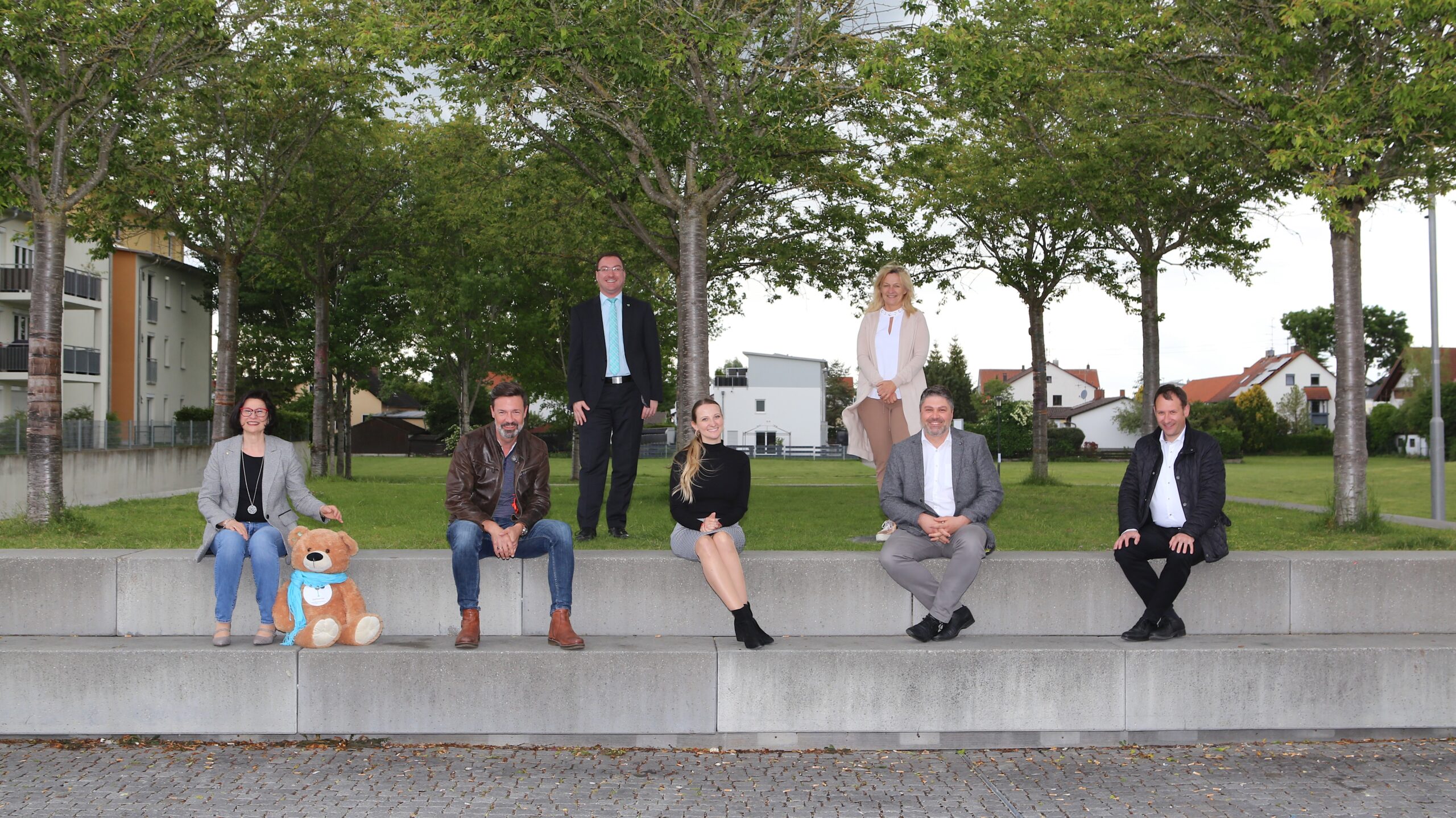 Gruppe von sieben Personen im Park auf Betonstufen, im Hintergrund Bäume und Gebäude; Aktionstag-Szene mit formeller und legerer Kleidung, MABP Umfeld.