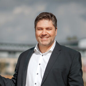 Alexnder Mademann in einem Anzug mit einem Lächeln, während er in die Kamera schaut.