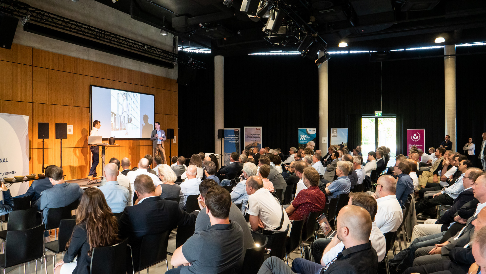 Blick auf die Konferenzbühne beim Commendit Unternehmerforum; zwei Redner präsentieren vor einem großen Publikum in einem Veranstaltungsraum