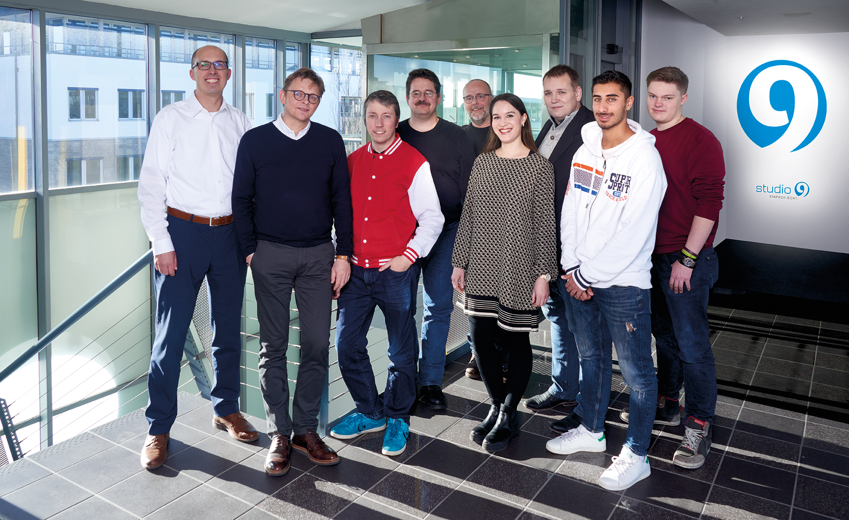 Gruppe von Mitarbeitern in einer modernen Innenraumlobby des Munich Airport Business Park, rechts Studio 9 Logo sichtbar, bunt gekleidet, Glass railing im Vordergrund