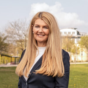 Doris Thalmeier mit langen blonden Haaren, trägt einen Blazer und lächelt in die Kamera.