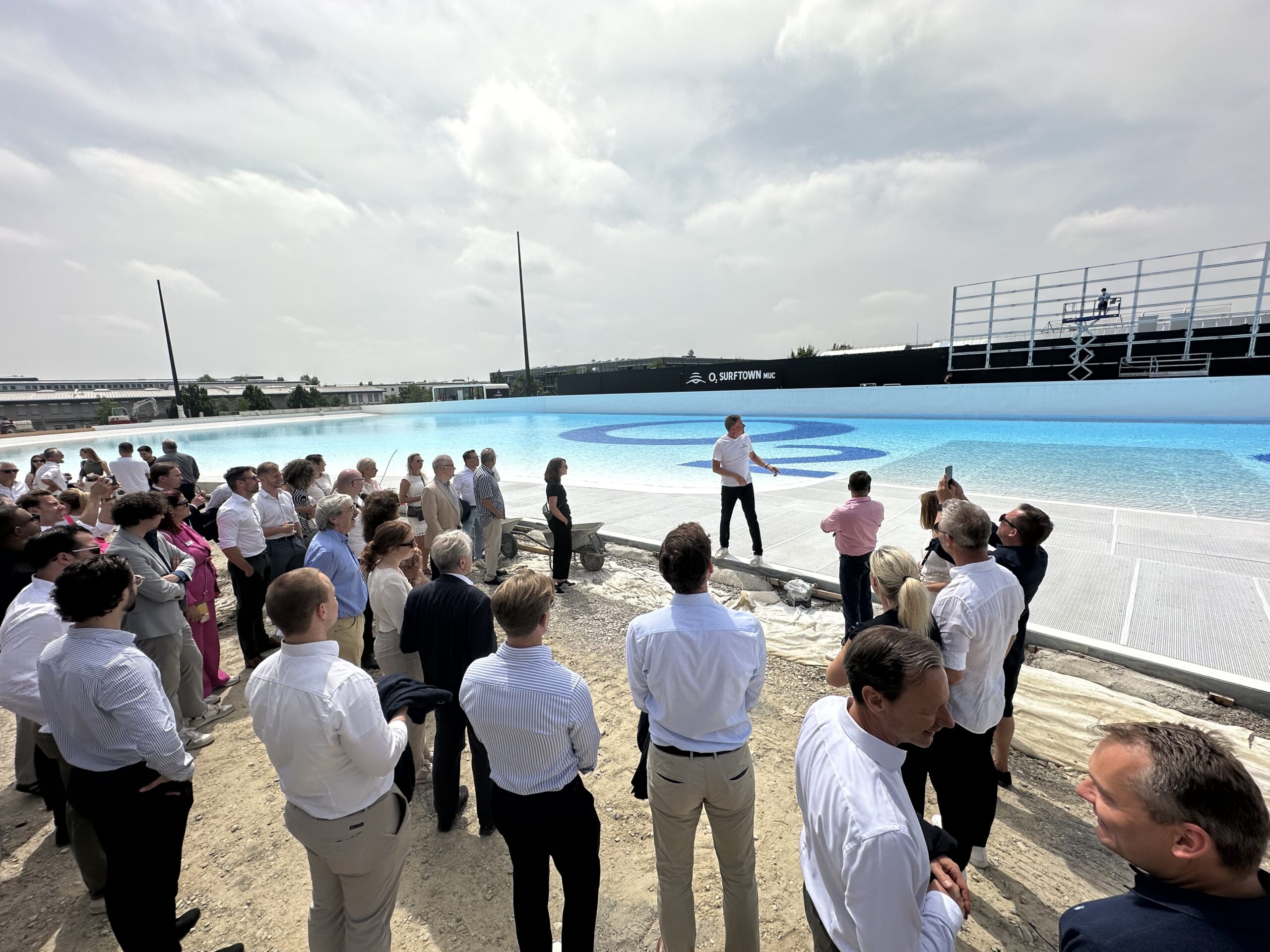 Eine Gruppe von Menschen hört einem Redner am Rand eines großen Pools zu, der mit Wasser gefüllt ist.