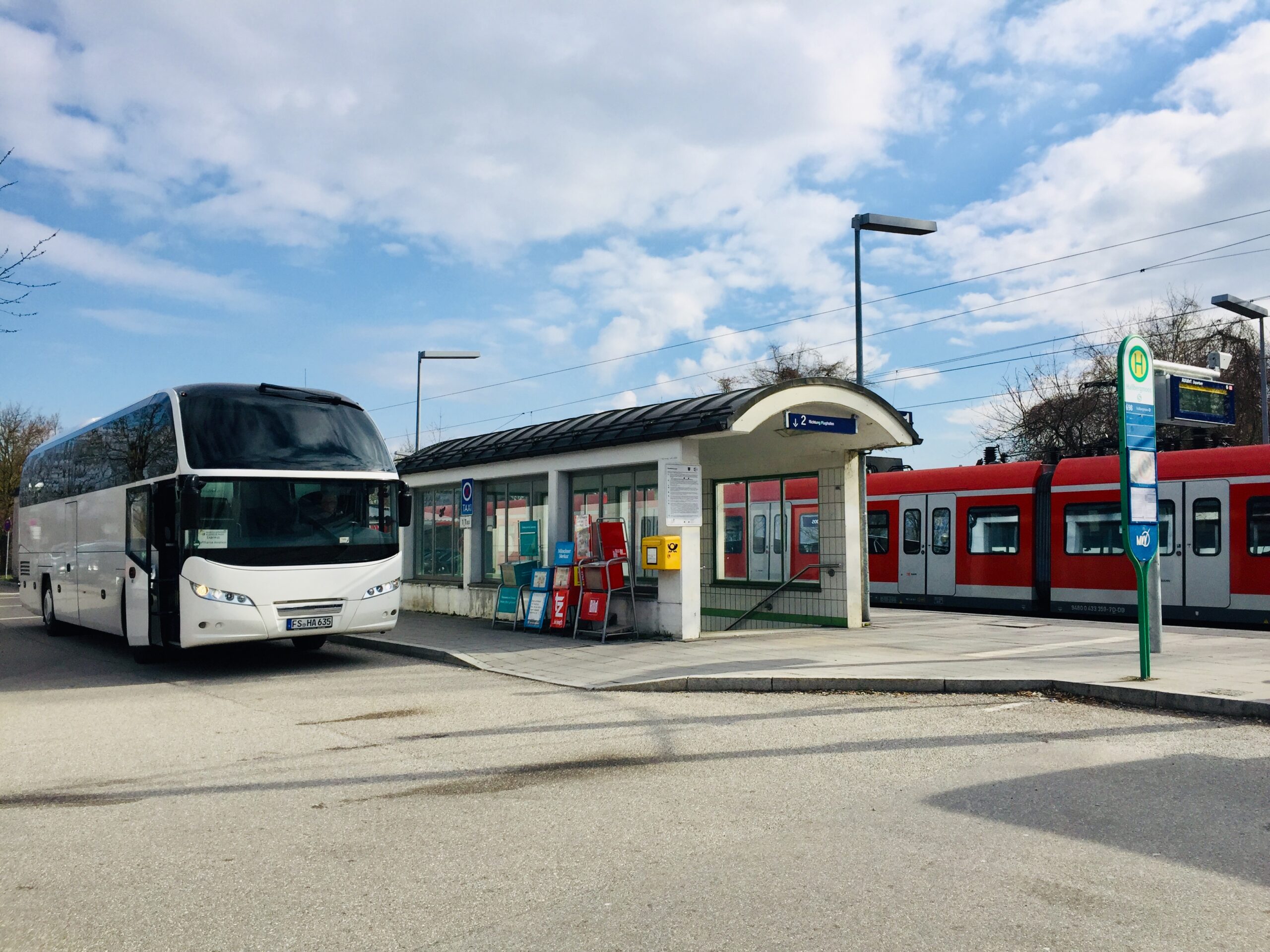 Weiße Reisebus am Haltepunkt neben einem roten Regionalzug, klarer Himmel über dem Bahnhofsbetrieb; Foto aufgenommen im Umfeld des MABP Munich Airport Business Park.