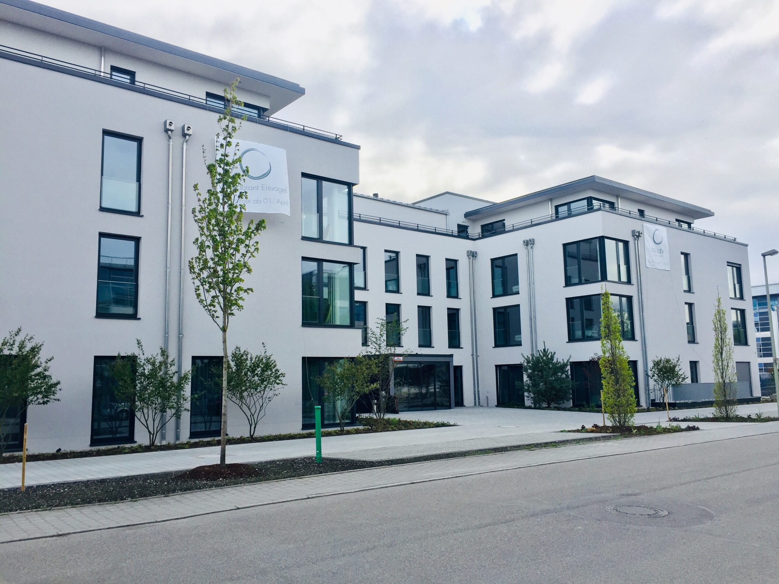 Mehrstöckiges weißes Bürogebäude im MABP – München Flughafen Business Park, moderne Fassade mit großen Fensterfronten, begrünter Vorhof und Parkplatzbereich
