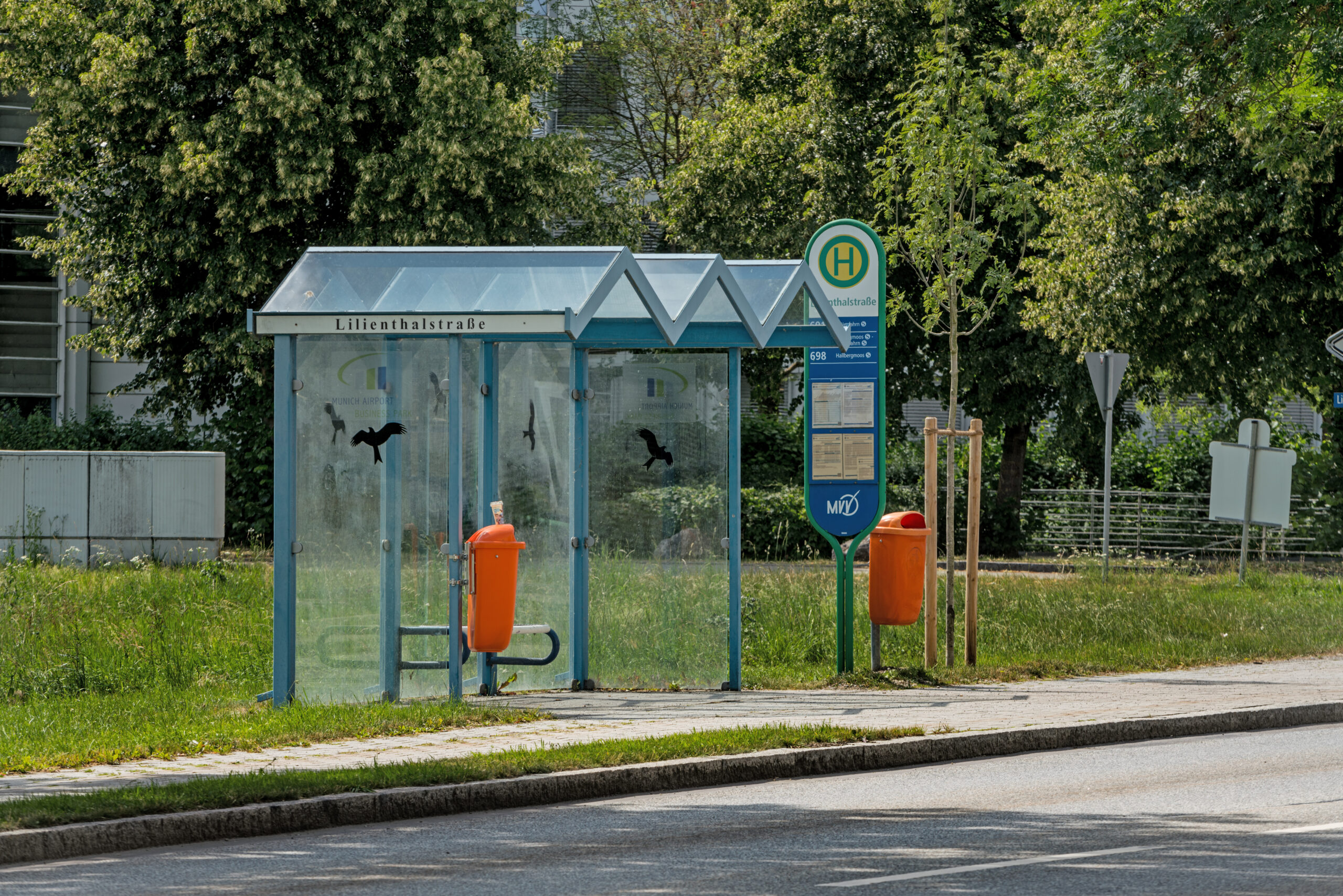 MABP Bus-Haltestelle Lilienthalstraße mit blauem Glas-Wartehäuschen, orangefarbenem Abfalleimer und grünem Umfeld am Straßenrand