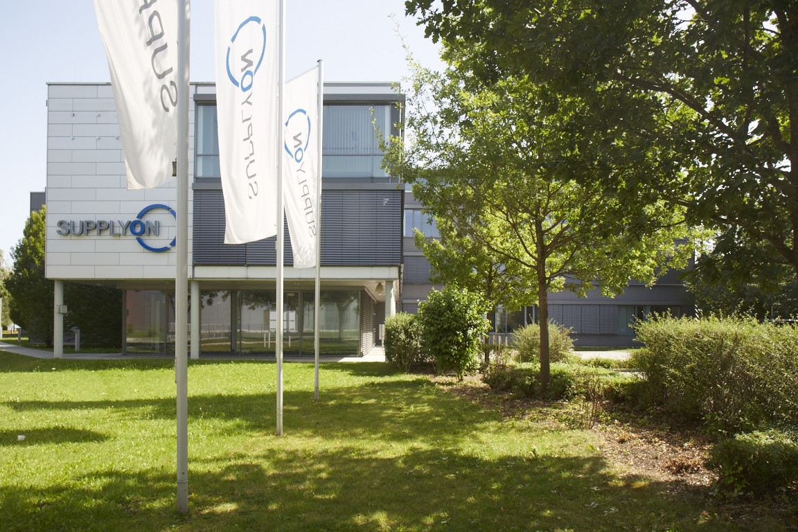 SupplyOn1-1: View of a modern office building in the Munich Airport Business Park, flanked by white flags, a green front garden, trees and shadows.