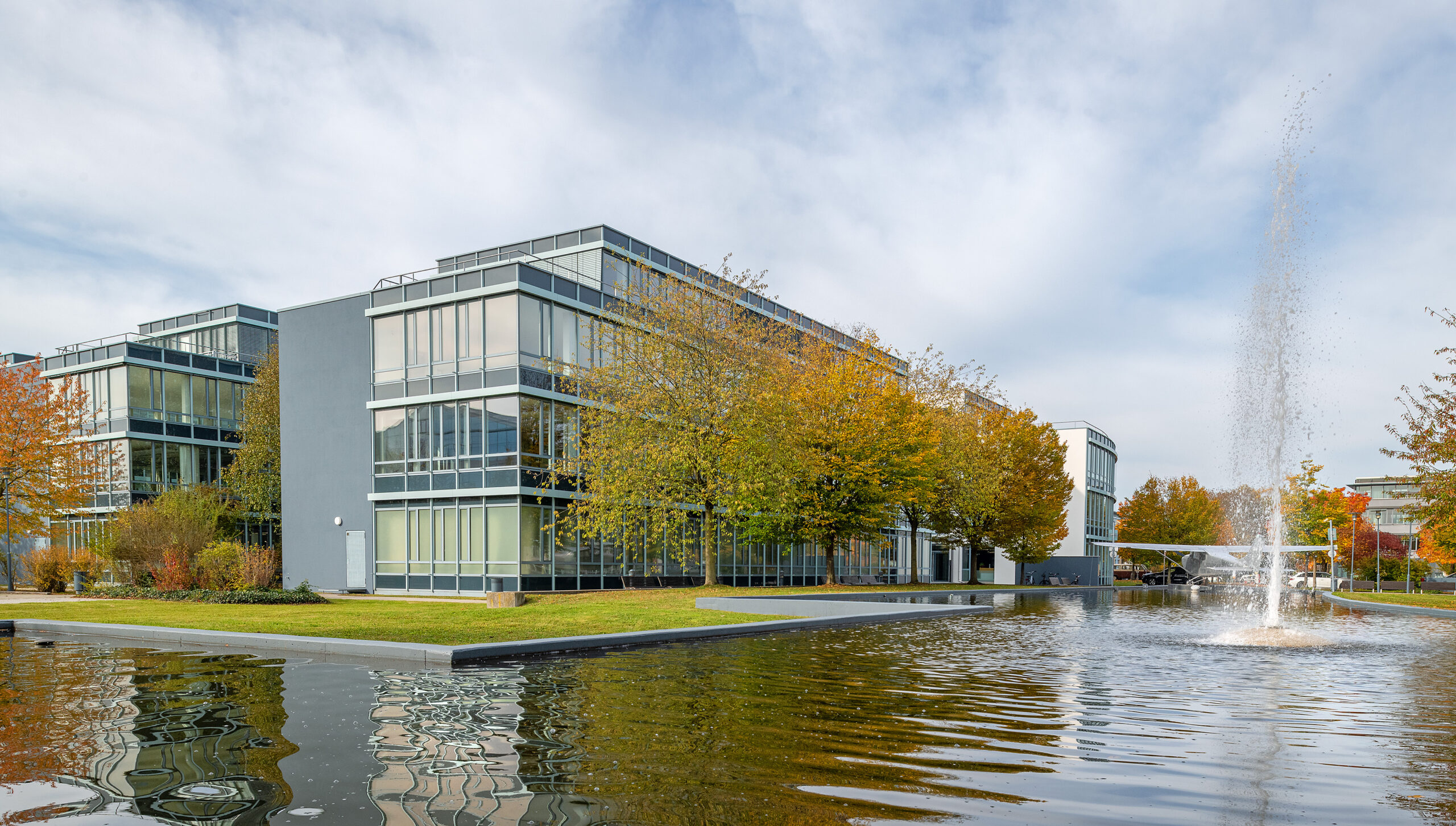 Architektur im Munich Airport Business Park mit modernem Fensterbau, Herbstbäumen in Orange- und Gelbtönen und einem großen Wasserbrunnen im Vordergrund, fotografiert von Claus Uhlendorf