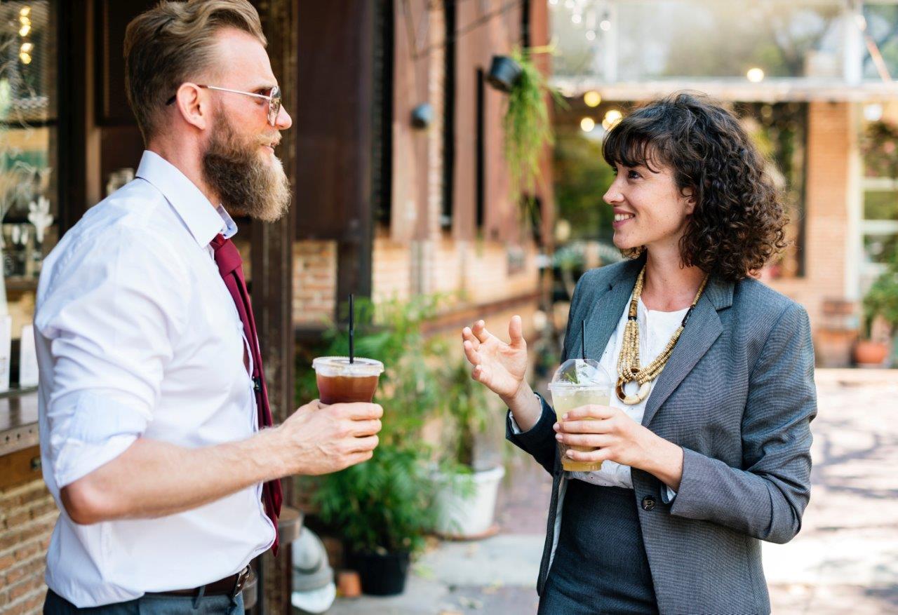 Two adults in business-casual style are standing outside in front of a café façade, holding transparent glasses with drinks, in conversation with each other.