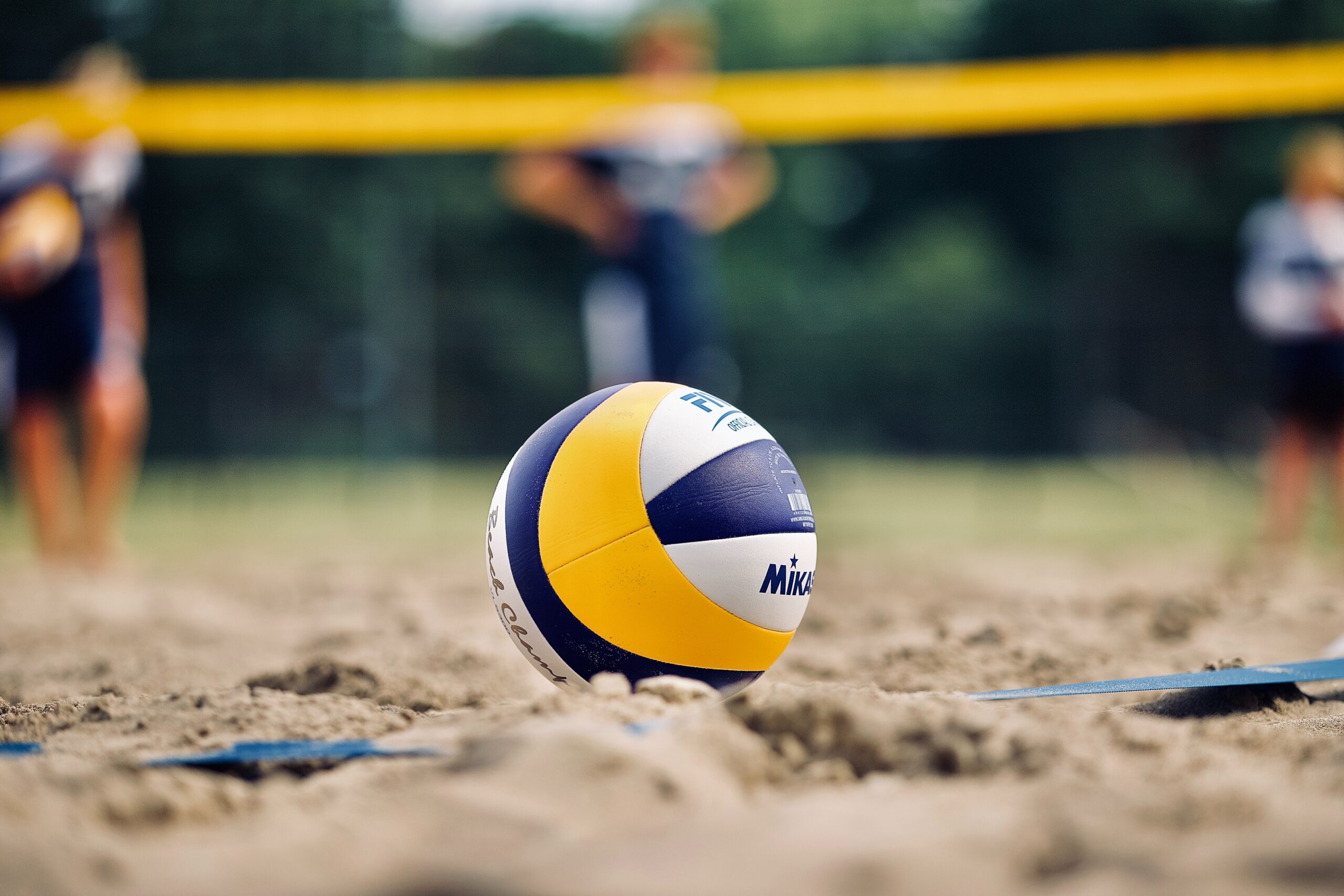 Miguel Teirlinck; gelb-blaues Volleyball auf Sandplatz, Fokus auf Ball, Netz im Hintergrund, unscharfe Gruppen in der Ferne, Bild von MABP Munich Airport Business Park