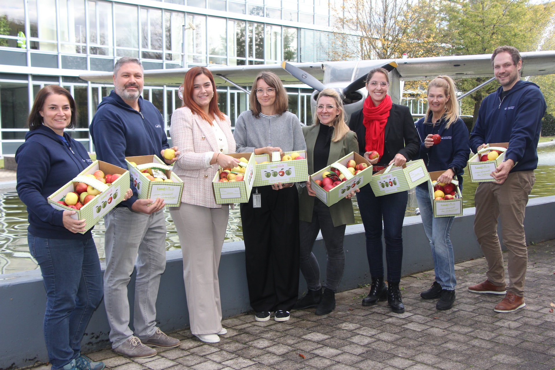 Gruppierung von Personen, die Apfelkisten in der Hand halten, vor einem modernen Gebäude im Freien.