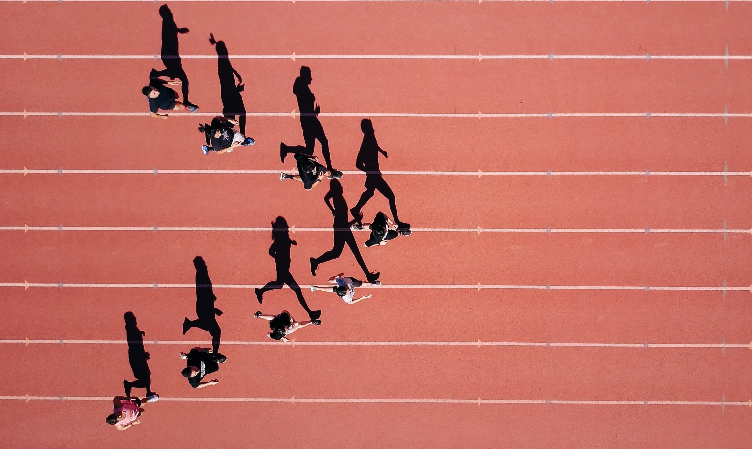 Steven Lelham Foto von oben: eine rote Leichtathletikbahn mit silhoutten von Menschen in Bewegung, lange Schatten verlaufen über die Bahn.