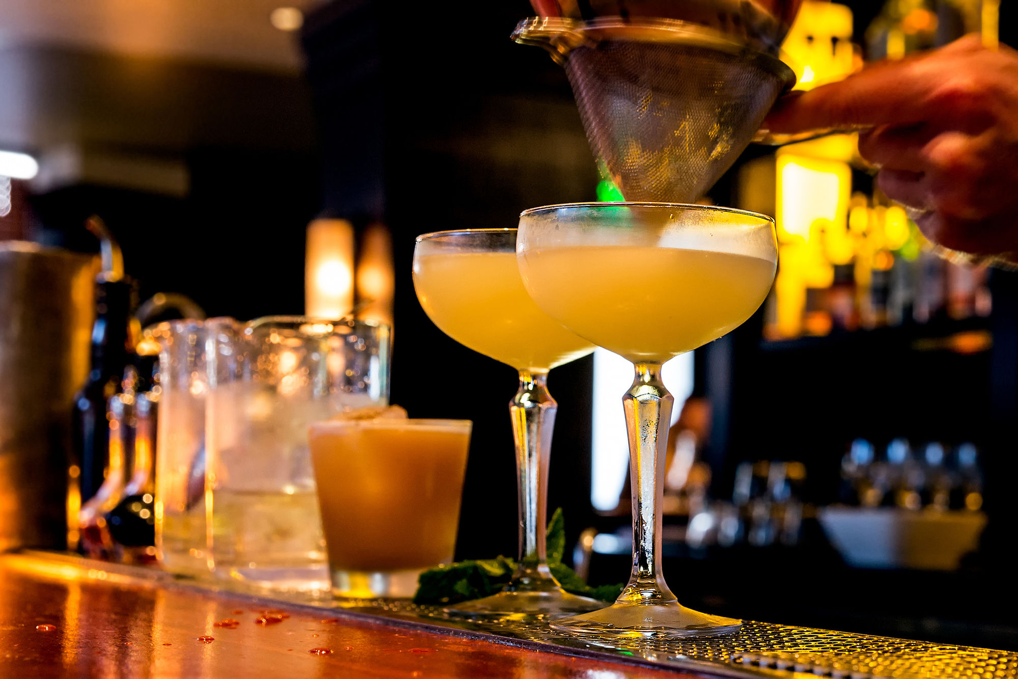 Zwei Cocktails in eleganten Gläsern, während ein Barkeeper einen Cocktail abseihen.