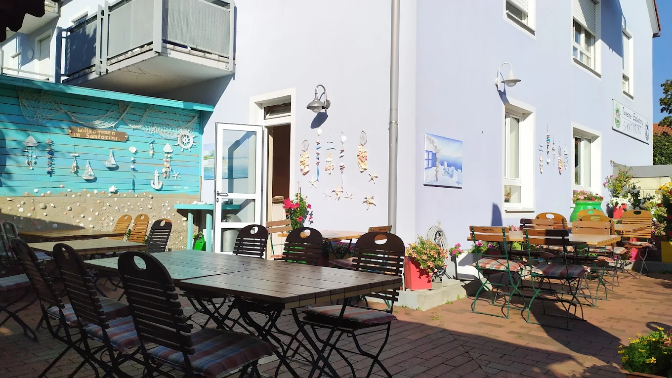 Tische und Stühle im Freien vor einem hellblauen Gebäude mit Dekorationen und Pflanzen.