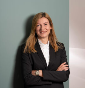 Portrait von Claudia-Zoric im SKYGATE: schwarzer Blazer, weiße Rüschenbluse, blondes Haar, Arme verschränkt; ruhiger Blick vor hellen Flächen.