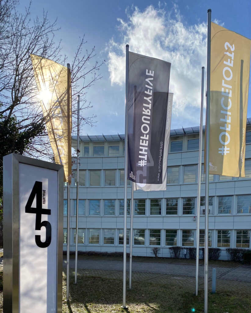 Außenansicht des Bürogebäudes im Munich Airport Business Park mit Flaggen, darunter gelbe und graue Fahnen, im Hintergrund blauer Himmel und die Bezeichnung FOURTYFIVE sichtbar