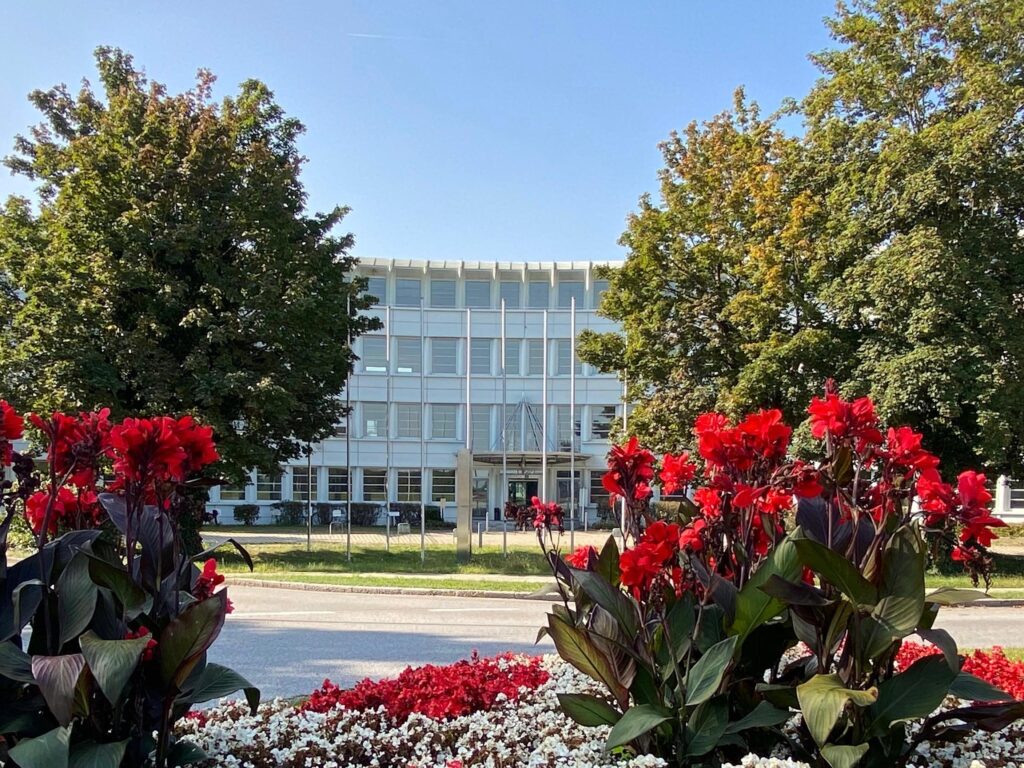 FourtyFive Büroloft-Gebäude im Munich Airport Business Park, umgeben von großen Bäumen und roten Blumen, moderne Architektur, flexible Büroflächen, Nähe zum Flughafen München