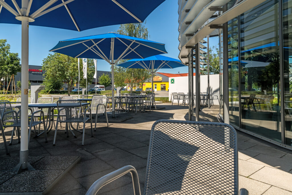 Tische und Stühle unter blauen Sonnenschirmen in einem Außenbereich eines Restaurants oder Cafés.