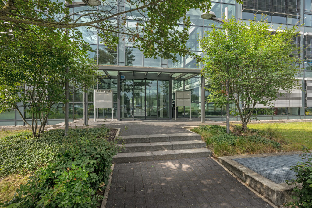 Eingang zu einem modernen Bürogebäude mit Glasfassade und begrünten Bereichen.
