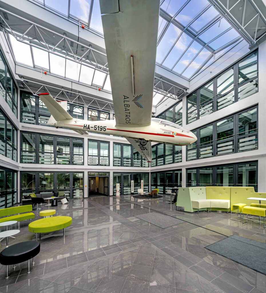 Ein Modell des Albatros-Hangars schwebt in einem modernen Innenraum mit großen Fenstern und Sitzgelegenheiten.