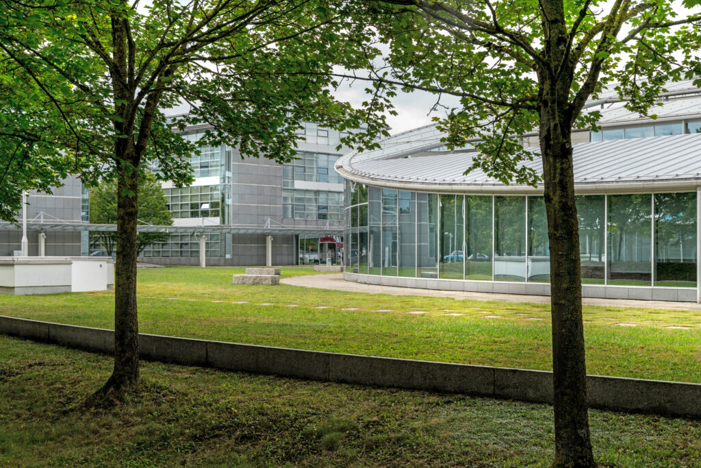 Moderne Gebäude mit großen Fenstern und einer grünen Rasenfläche im Vordergrund, umgeben von Bäumen.