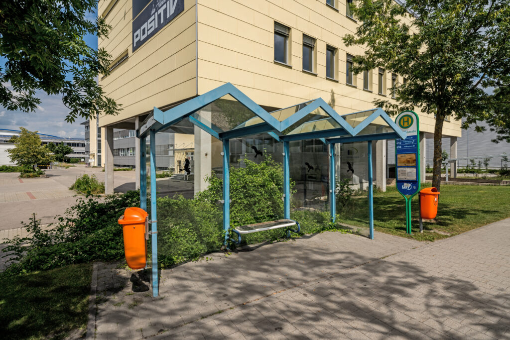 Haltestelle mit einem modernen, gläsernen Überdach und einer Bank in einem urbanen Umfeld.