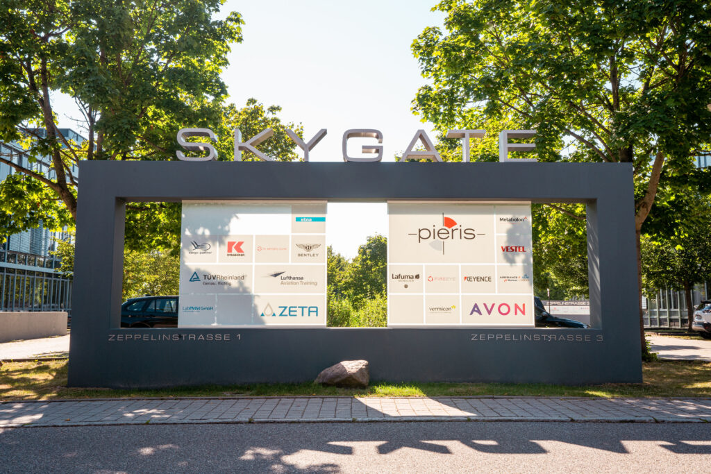 Skygate-Schild an der Zeppelinstraße mit Logos von Unternehmen wie Lufthansa, Nestlé und Avon.