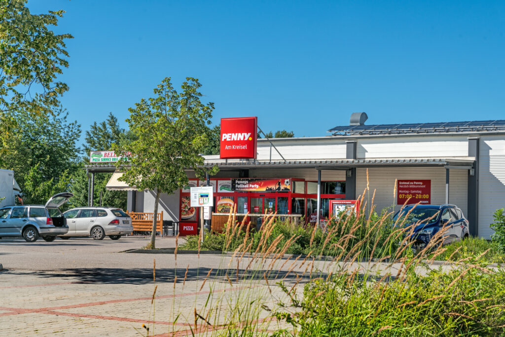 PENNY Supermarkt mit rotem Logo und Eingangsbereich, umgeben von Bäumen und Parkplätzen.