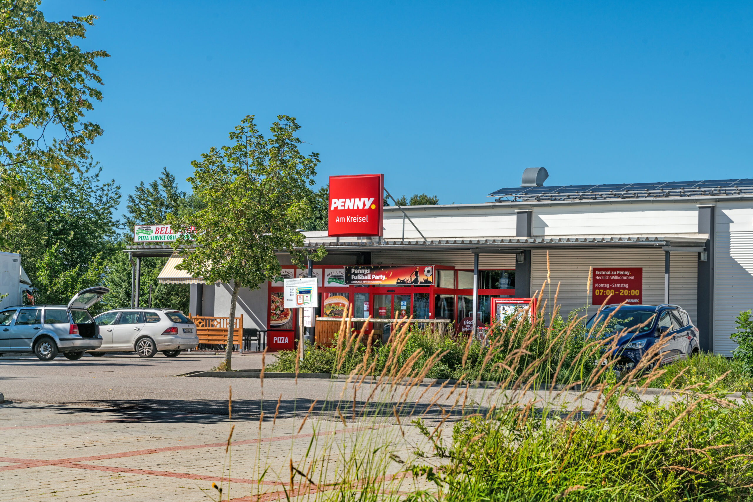 PENNY Supermarkt mit rotem Logo und Eingangsbereich, umgeben von Bäumen und Parkplätzen.