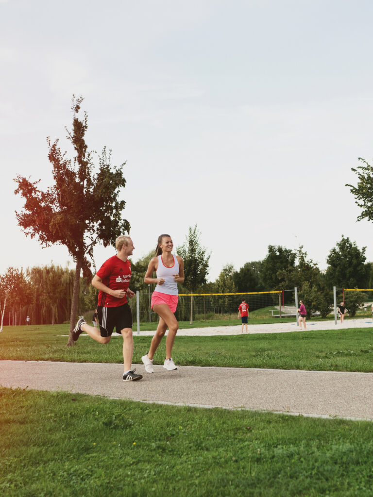 Zwei Personen joggen auf einem Weg in einem Park, umgeben von Bäumen und Sporteinrichtungen.